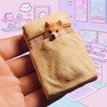 A small decorative figurine of a Shiba Inu dog peeking out from under a tan blanket in a miniature bed, held in a human hand.