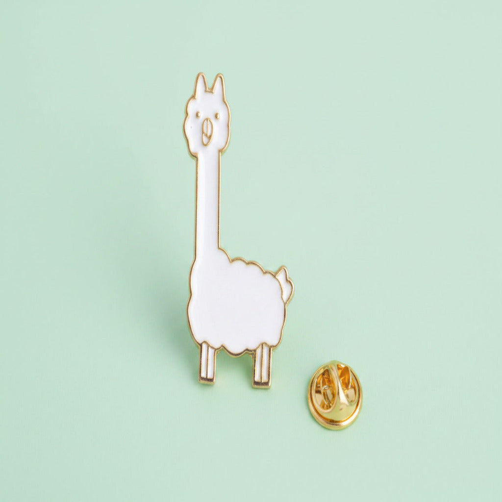 A white llama enamel pin with gold metal outlines, shown next to its gold pin backing on a plain white background.