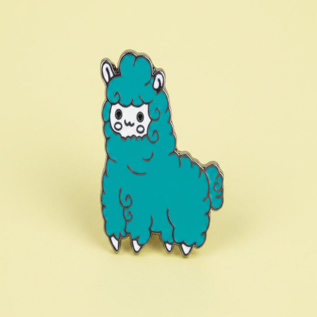 A cute alpaca enamel pin featuring a turquoise blue body, white face and hooves, and silver metal outlines, shown on a pastel yellow background.