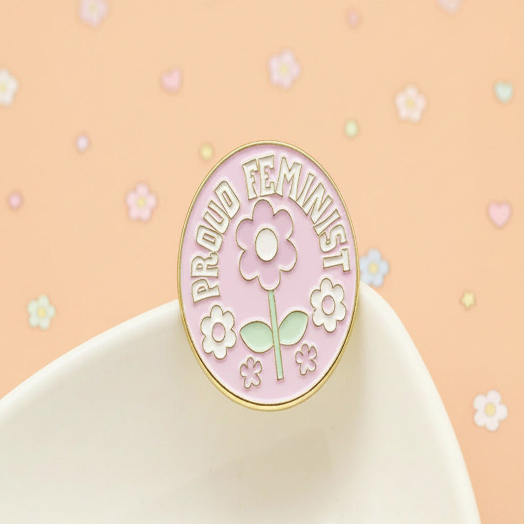A round enamel pin with a pink background, gold metal border, and a central flower design with the words 'PROUD FEMINIST' in white. The pin is displayed on a white ceramic dish against a soft yellow background.