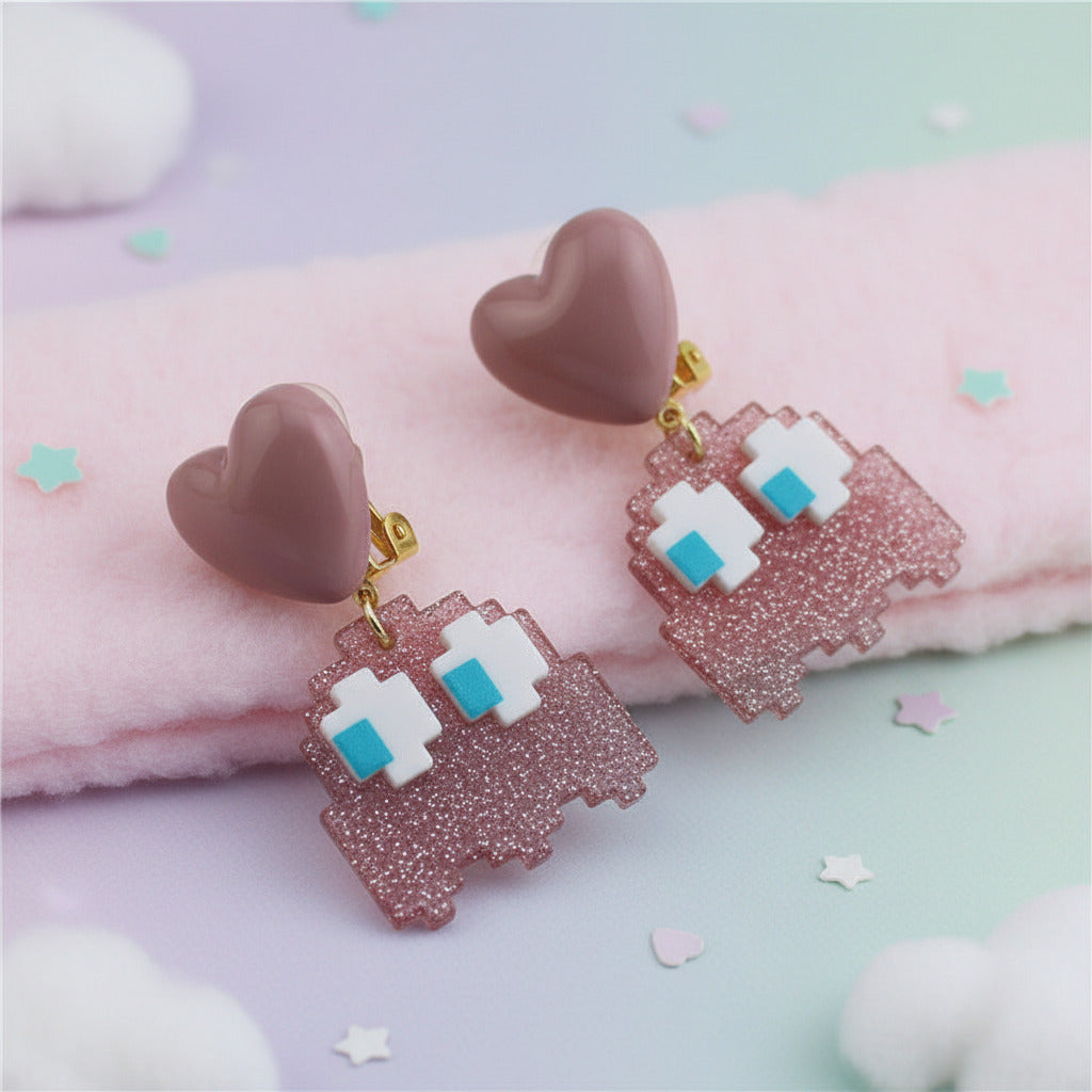 A pair of pink and gold clip-on earrings displayed on a light gray marble hexagonal tray. One earring features a solid dusty pink heart-shaped stud with a gold clip, while the other features a glittery pink ghost-shaped drop with white pixel eyes and small blue accents, attached to a pink heart stud.