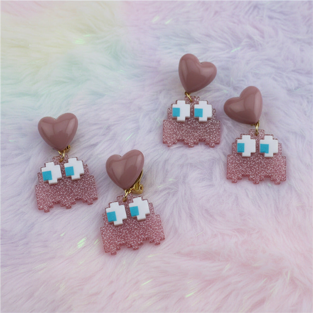 Four matching dangle earrings laid on a light gray textured surface. Each earring has a dusty pink heart-shaped top, a pink glitter middle section, and a dangling pink glitter ghost charm with white pixel eyes and small turquoise blue accents.