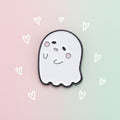 A cute cartoon ghost charm or pin with a white face, black outline, and pink cheeks on a pastel colored background.