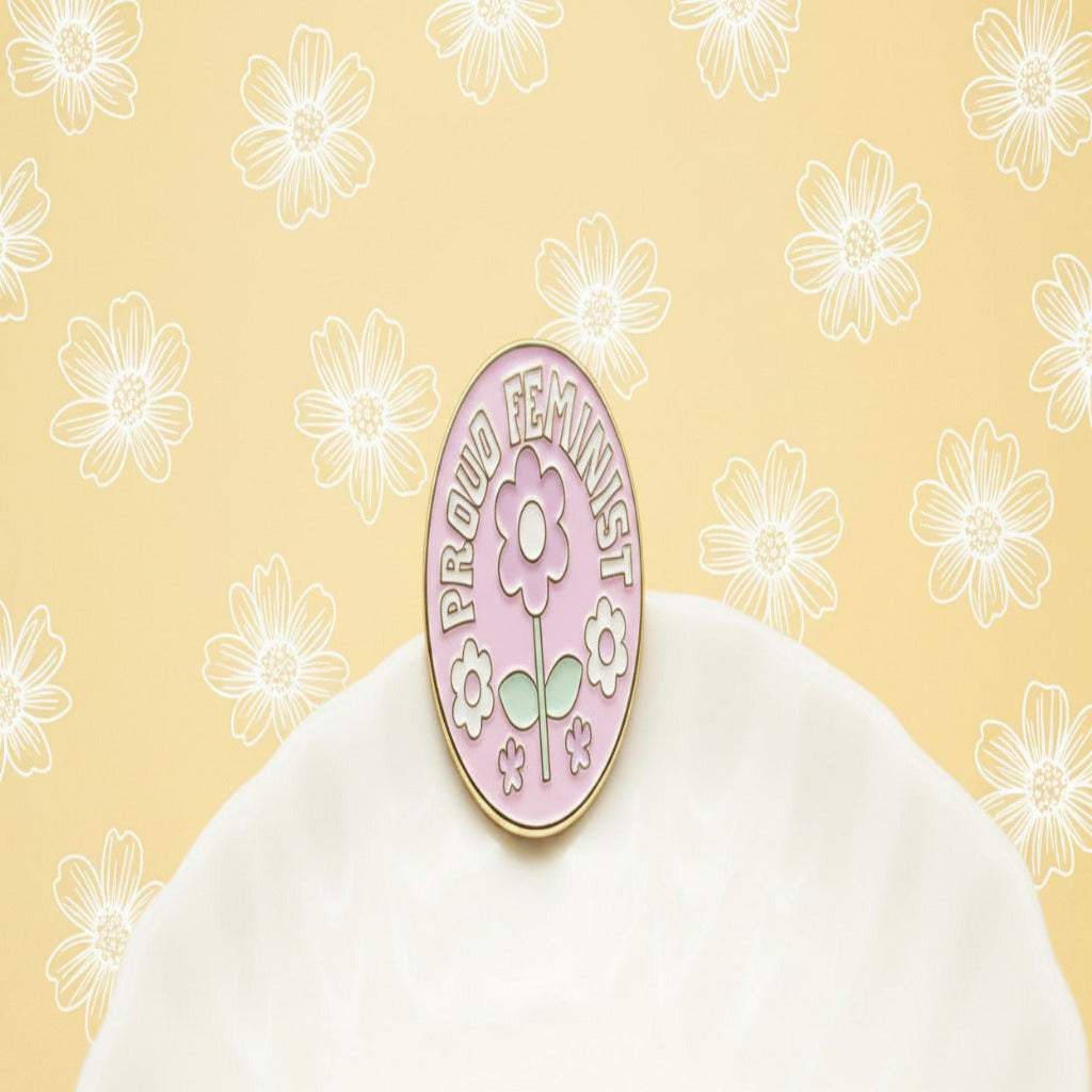 Two round enamel pins displayed on a white surface. One pin is light pink with a white flower, green stem, and the words 'PROUD FEMINIST' in white.