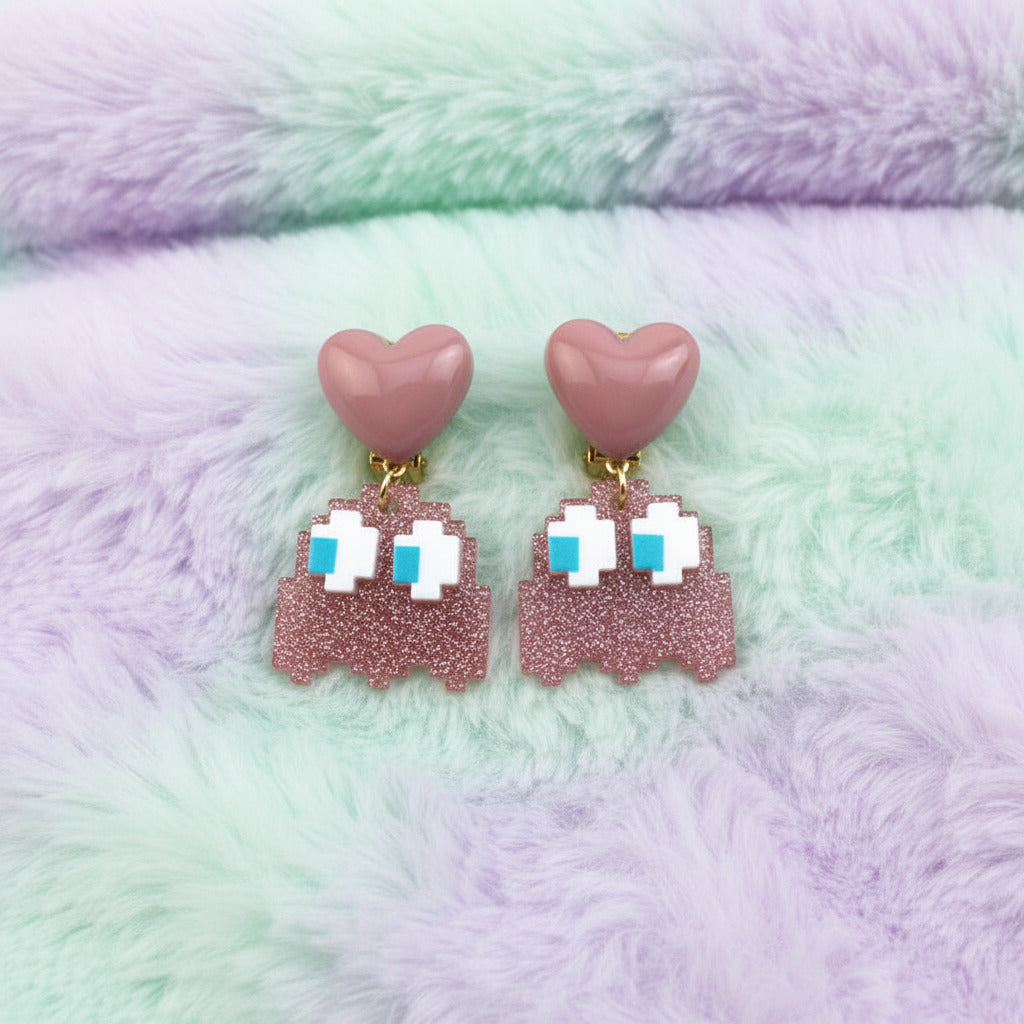 A pair of dangling earrings featuring pink heart-shaped studs and pixelated ghost charms in a glittery pink, white, and light blue design, shown on a plain white background.