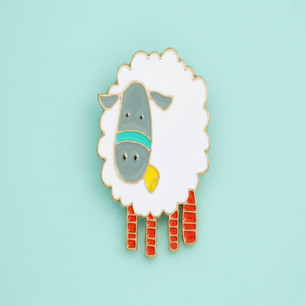 A cute cartoon sheep enamel pin with a white woolly body, gray face, teal bandana, yellow bell, and red striped legs on a plain white background.