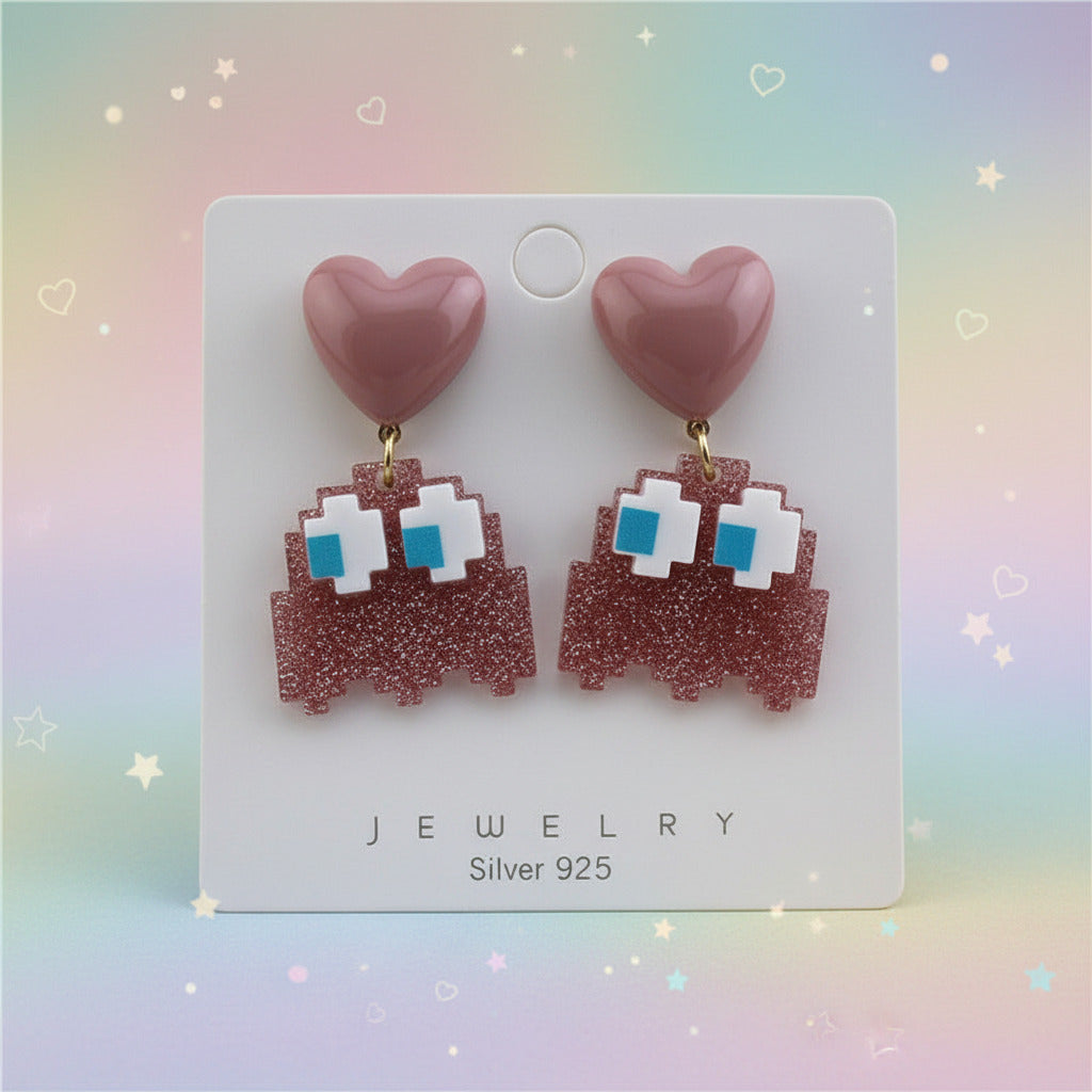 A pair of retro pixel-art dangling earrings displayed on a white jewelry card. Each earring has a dusty pink heart-shaped top with a dangling glittery pink pixelated body featuring white and bright blue square details, suspended by small gold rings.