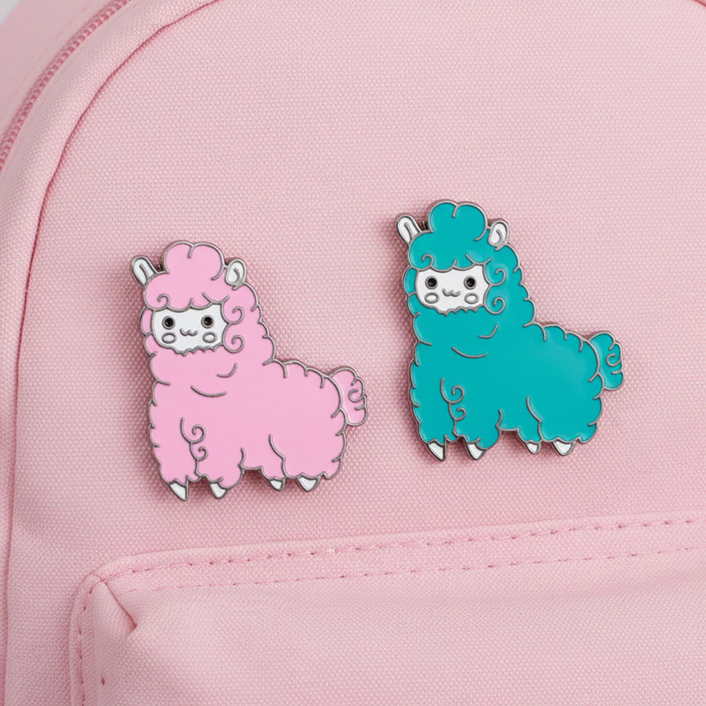 Two cute alpaca enamel pins, one pink and one teal, displayed on a blue distressed pink canvas background.
