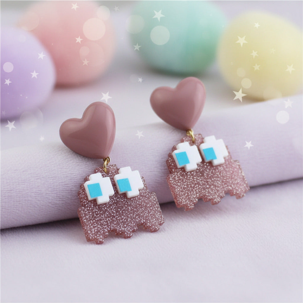 A pair of dangling earrings displayed on a dark stone slab. Each earring has a dusty pink heart-shaped top, a glittery pink pixelated ghost-like body, and white square eyes with small blue centers.