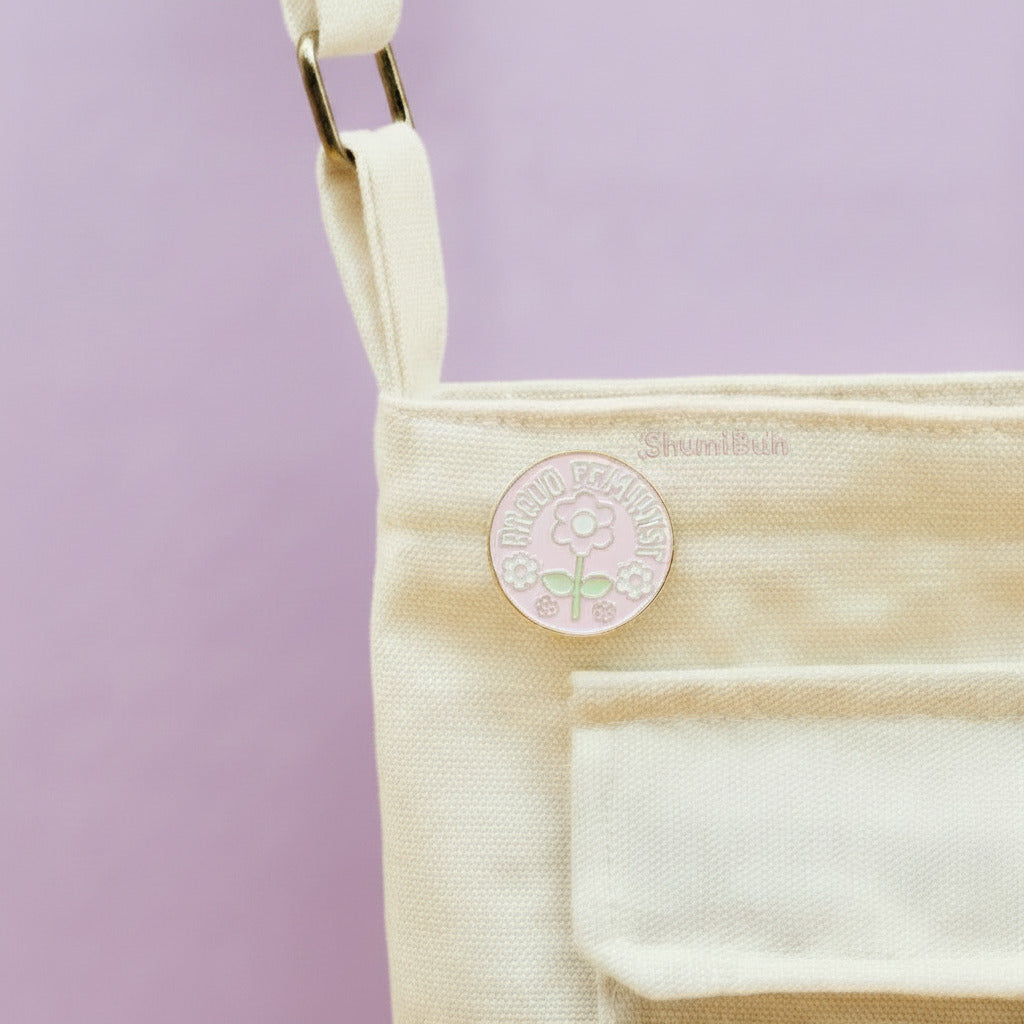 Close-up of a cream canvas shoulder bag with two round enamel pins attached to the front pocket, set against a solid yellow background.