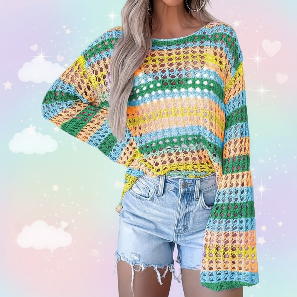 A woman wearing a colorful crocheted knit sweater with wide sleeves and horizontal stripes in green, blue, yellow, orange, and white, paired with light-wash denim shorts.