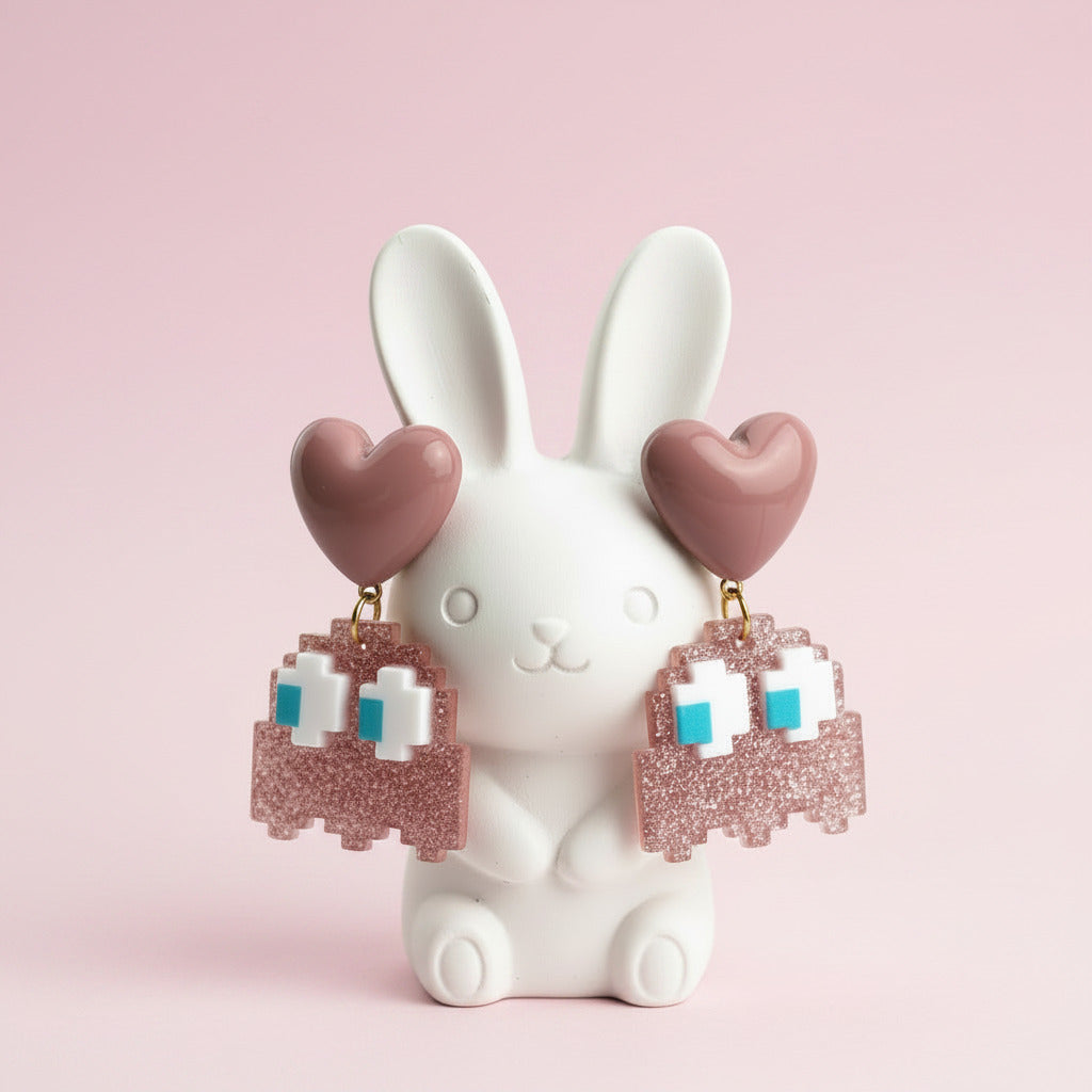 A pair of dangling earrings displayed on a black T-shaped stand against a white background. Each earring has a pink heart-shaped top and a glittery pink ghost-shaped dangle with white and light blue pixel details.