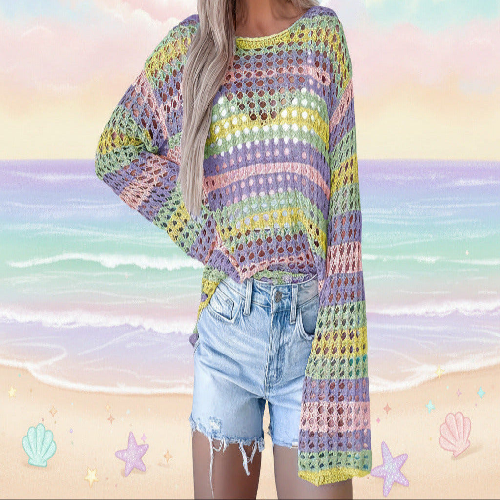 A woman wearing a colorful, open-knit crochet sweater with wide bell sleeves and light-wash denim shorts, standing against a white paneled wall.