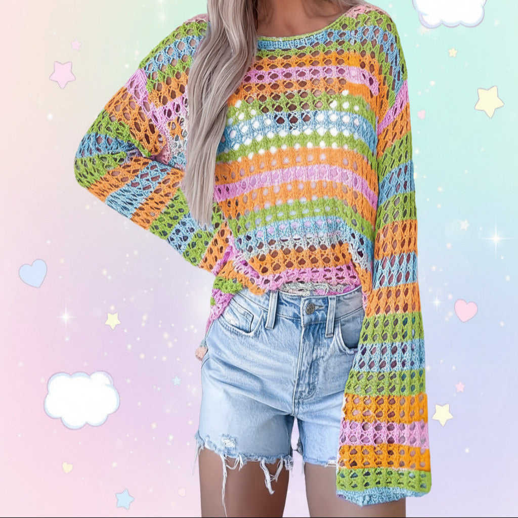 A woman wearing a colorful striped crochet knit sweater with wide bell sleeves and light-wash denim shorts, standing against a light gray paneled wall.