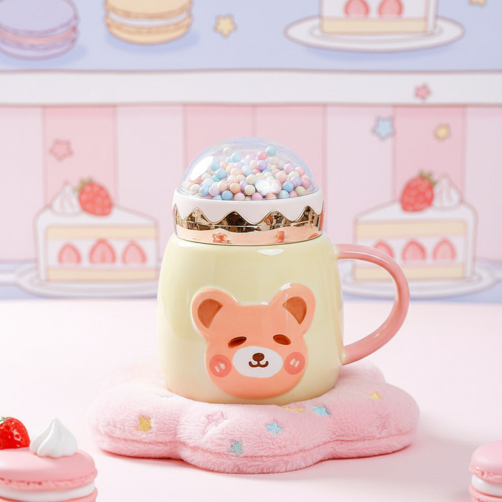 A cute ceramic mug with a bear face design and a pink handle, shown on a table with a person holding a book in the background. The mug has a gold rim and a clear dome lid filled with colorful beads.
