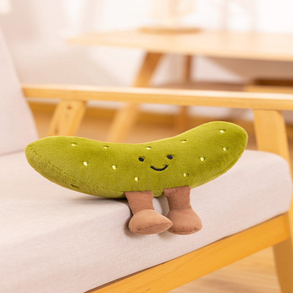 A green pickle-shaped plush cushion with a smiling face and small tan legs, resting on a light-colored chair in a softly blurred indoor setting.