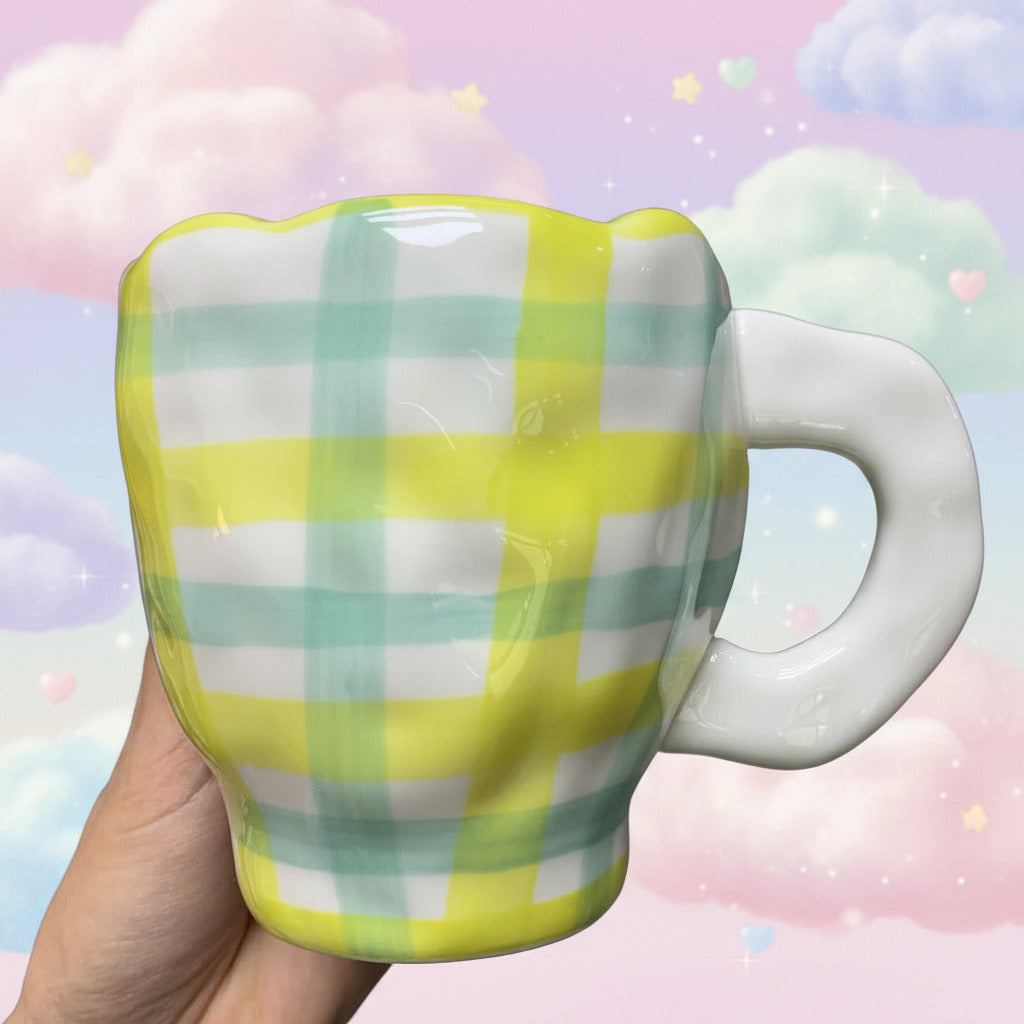 A hand holding a ceramic mug with a yellow, green, and white plaid pattern and a glossy white handle, set against a blurred gray background.