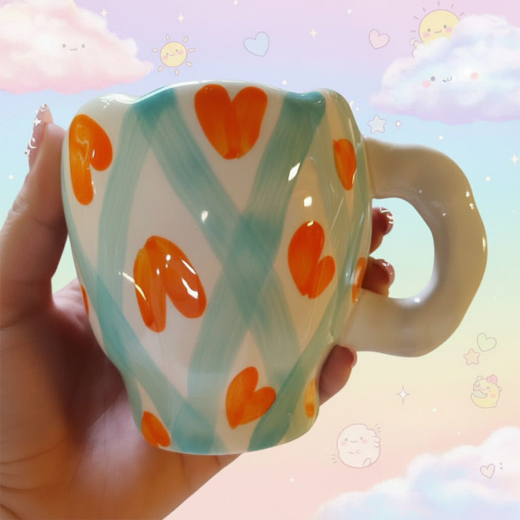 A hand holds a white ceramic mug decorated with painted light blue brushstroke patterns and orange heart motifs, against a blurred gray floor background.