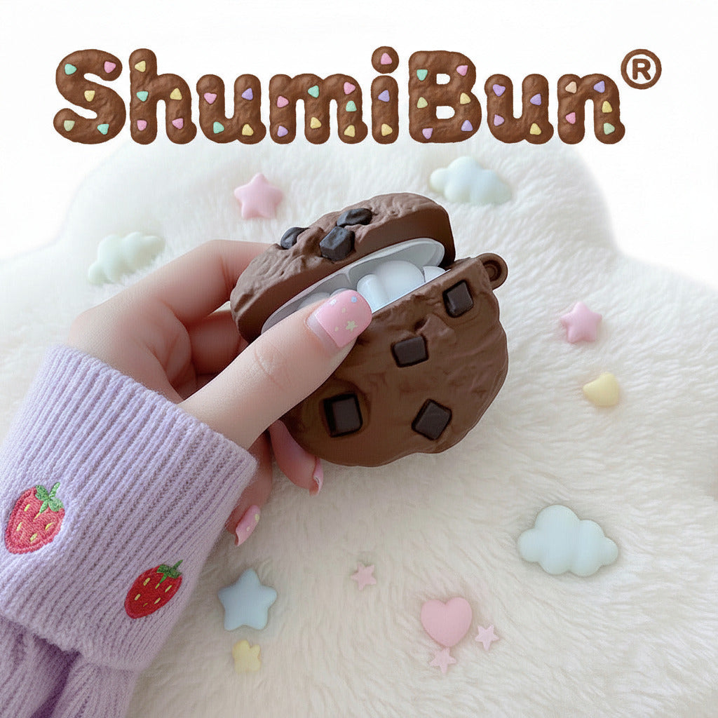 A hand wearing a gray sleeve holds a chocolate chip cookie-themed AirPods case, which is open to reveal white earbuds inside, set on a wooden table with a coffee cup and cookies in the background.