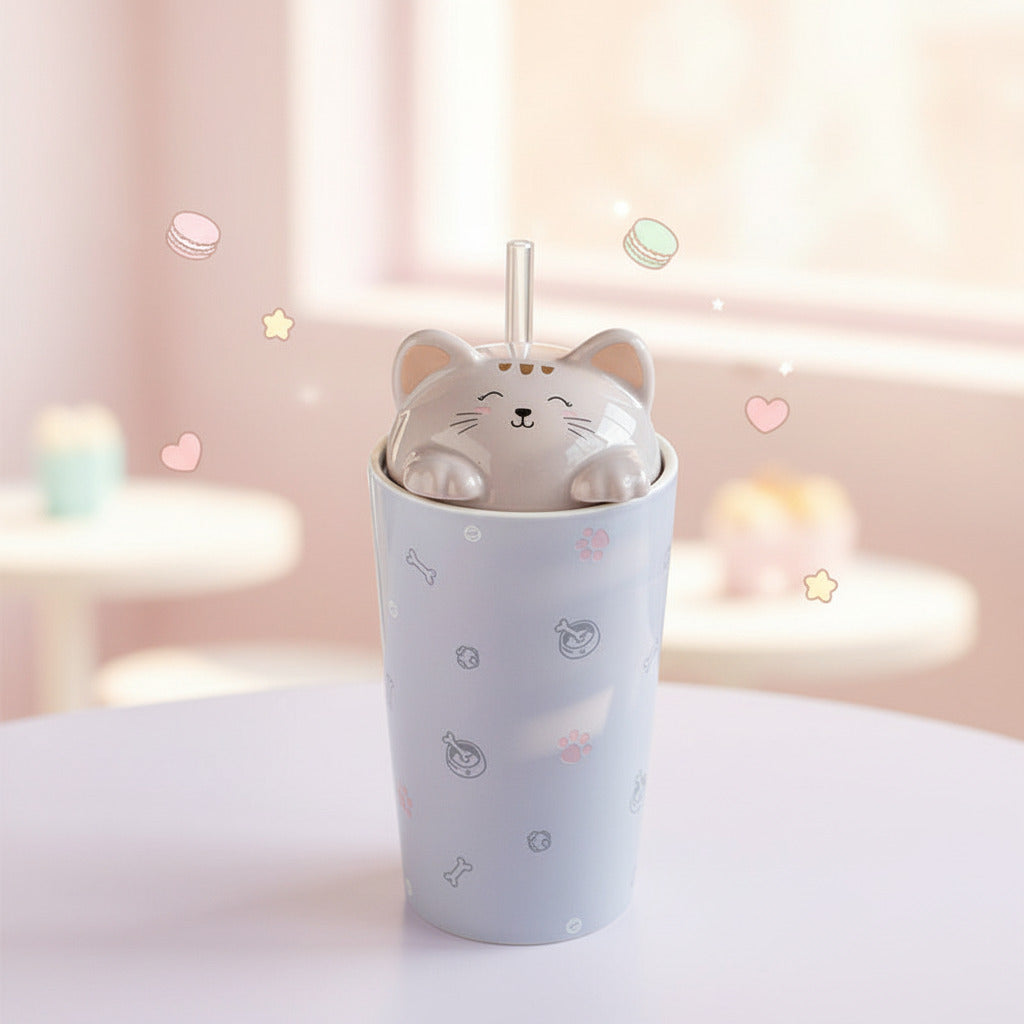A light lavender ceramic tumbler cup with a matching cat-shaped lid and straw, shown on a white tabletop with a blurred book and dried flowers in the background.