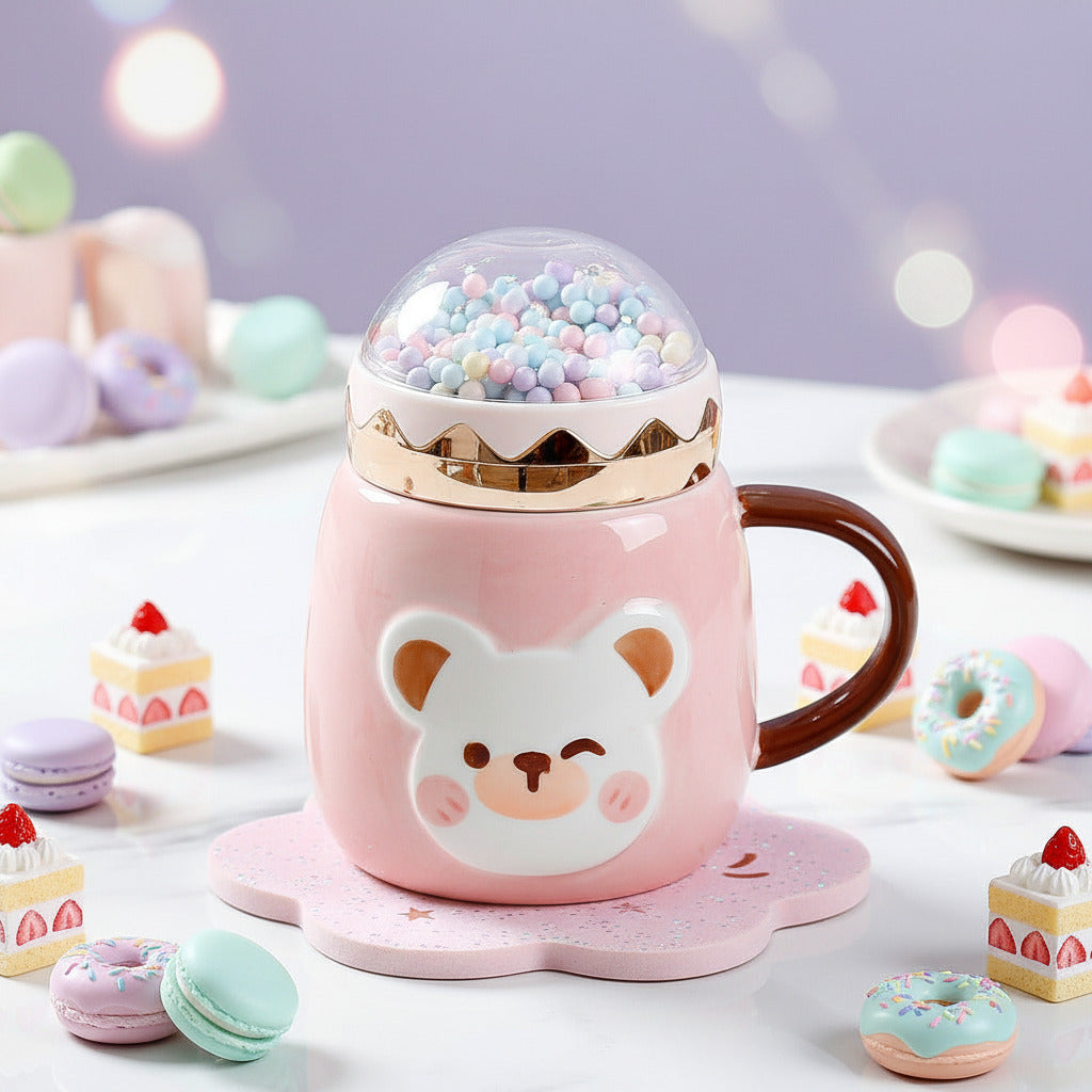 A pink ceramic mug with a bear face design, a gold rim, and a clear dome lid filled with pastel beads, shown on a tabletop with a coaster and coffee cup nearby.