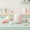 A pink ceramic mug with a white polar bear decoration and a white lid featuring a small bear-shaped spoon handle, shown on a light blue background with two macarons beside it.