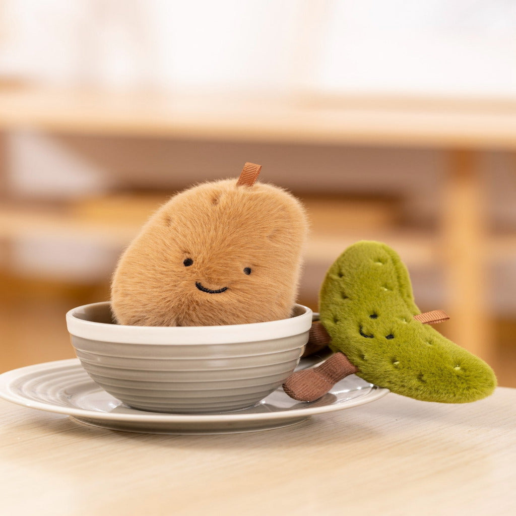 A plush toy shaped like a smiling beige pear or fruit sits in a small white cup on a saucer, with a green leaf-shaped plush arm resting beside it on a wooden table.