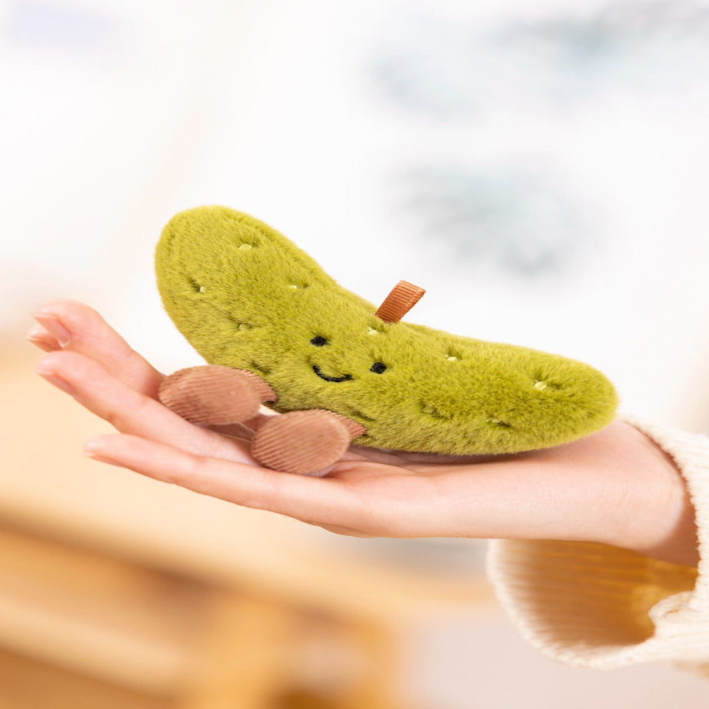 A small plush pickle-shaped toy with a smiling face and brown feet is held in a person's hand. The toy has a fuzzy olive-green body with embroidered details and a small brown stem, set against a softly blurred indoor background.