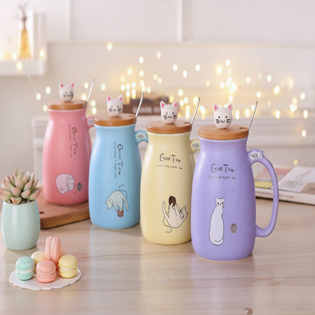 Four ceramic mugs with wooden lids and metal spoons, decorated with cute cat illustrations and the text 'Good Time'. The mugs are in pink, light blue, pale yellow, and lavender, arranged on a wooden table with a blurred background of flowers and books.