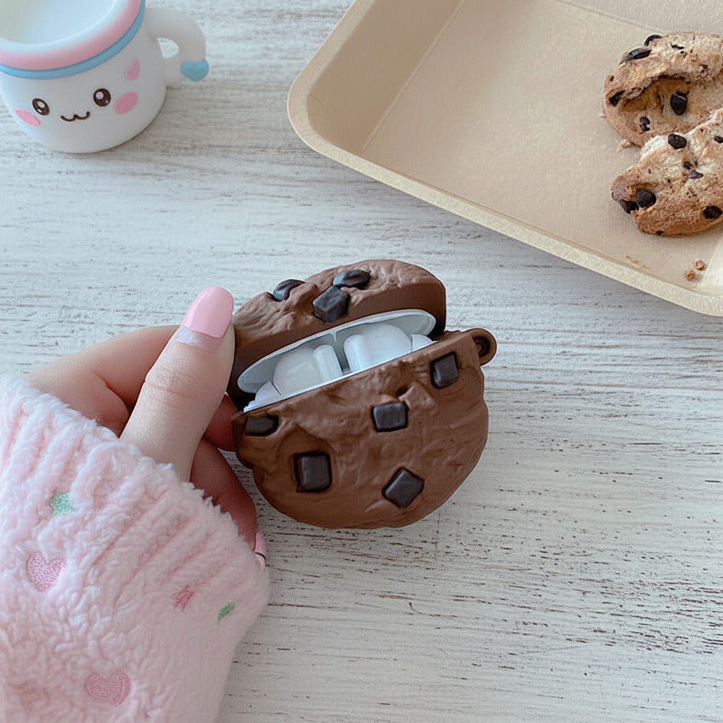A hand wearing a pink sleeve holds a chocolate chip cookie-themed AirPods case, which is open to reveal white earbuds inside, set on a wooden table with a coffee cup and cookies in the background.