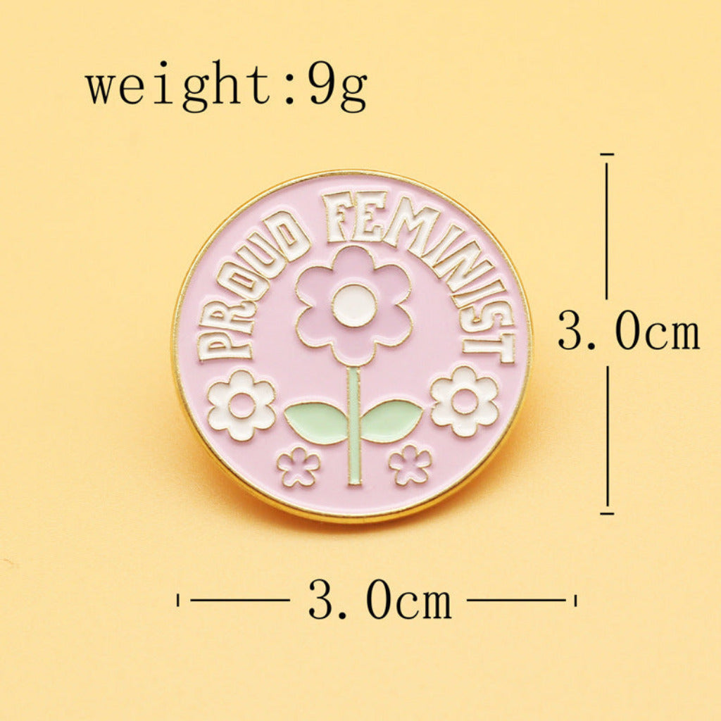 A round enamel pin with a pink background, white text reading 'PROUD FEMINIST,' and white flower outlines with pale green leaves. The image includes dimension lines and text showing the pin is 3.